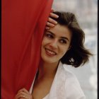 Woman with dark hair and white blouse peeking from red curtain