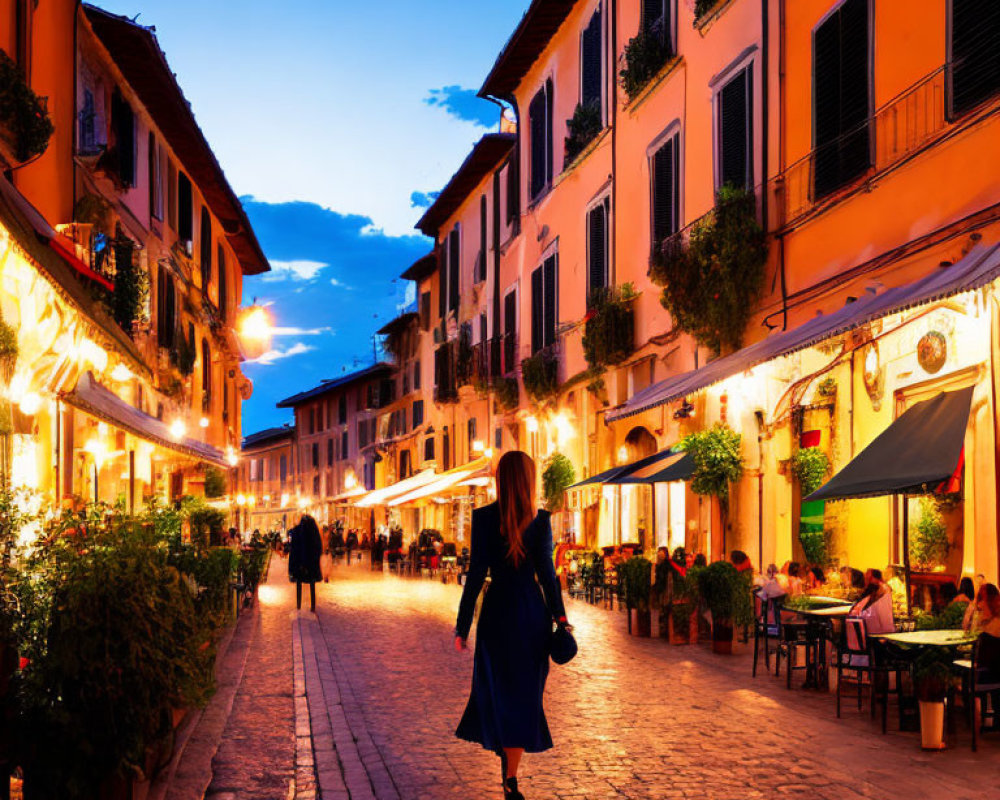 Woman in Blue Dress Walking on Illuminated Cobblestone Street at Dusk