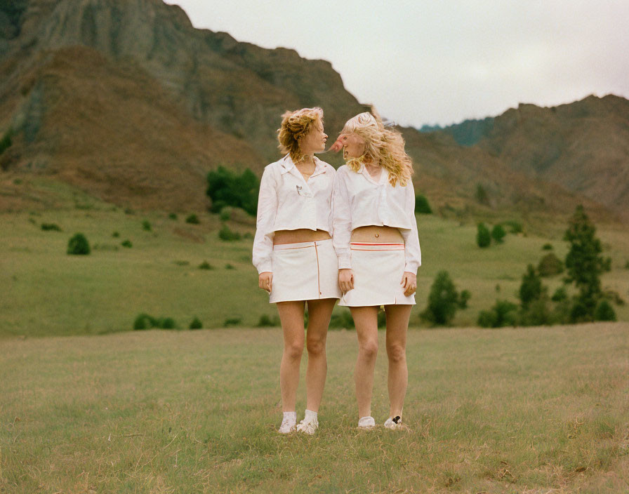 Two women in white standing back to back in grassy field with hills.