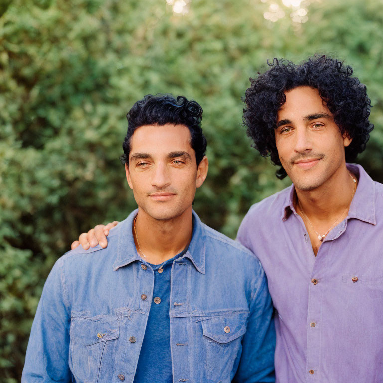 Two Men with Curly Hair in Blue and Purple Shirts Against Green Leafy Background