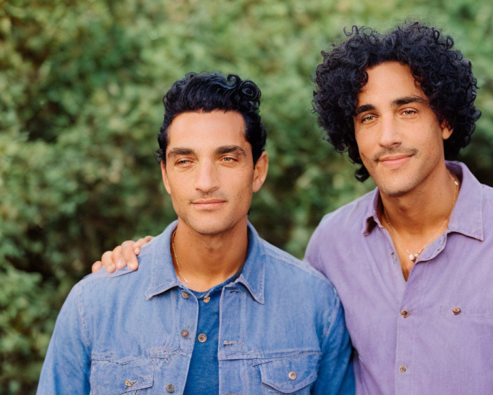 Two Men with Curly Hair in Blue and Purple Shirts Against Green Leafy Background