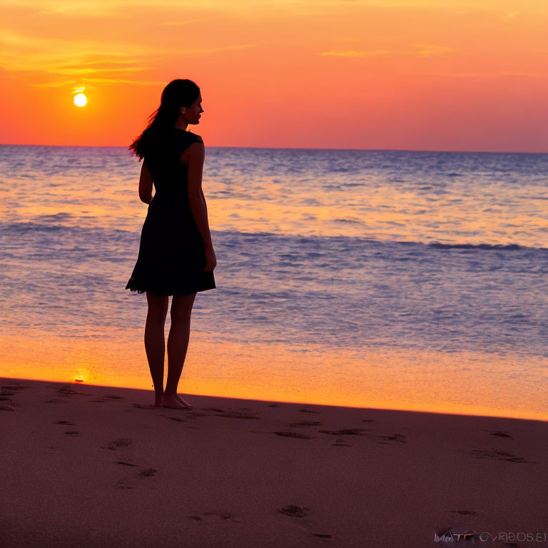 Woman silhouette standing on beach at sunset with low sun and water reflection