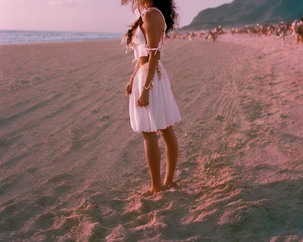 Woman in white dress on sandy beach at dusk barefoot, hair blowing.