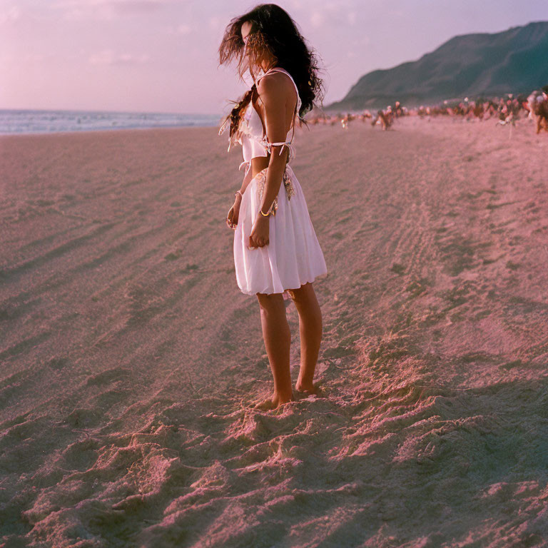 Woman in white dress on sandy beach at dusk barefoot, hair blowing.