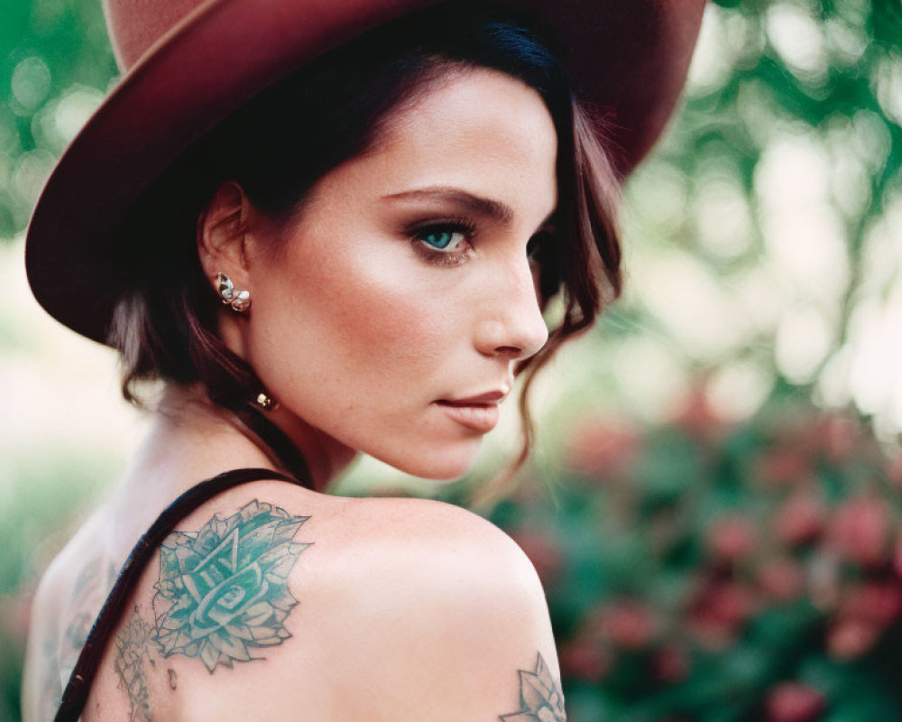 Woman with shoulder tattoo wearing hat, blurred greenery and flowers background