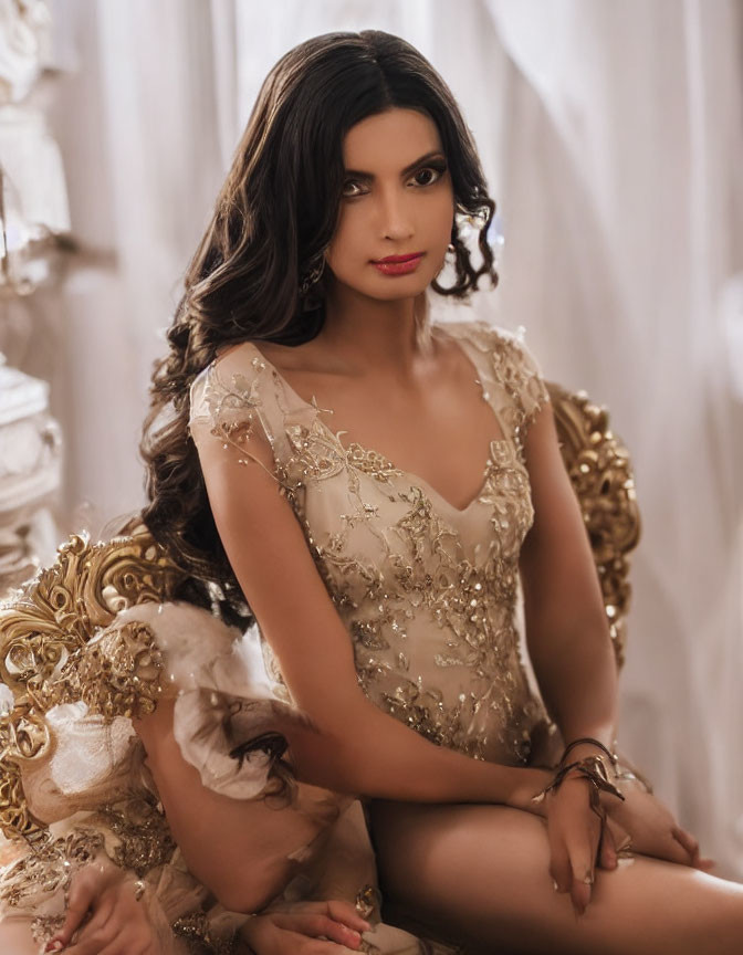 Dark-haired woman in gold dress sitting on ornate chair gazes serenely.