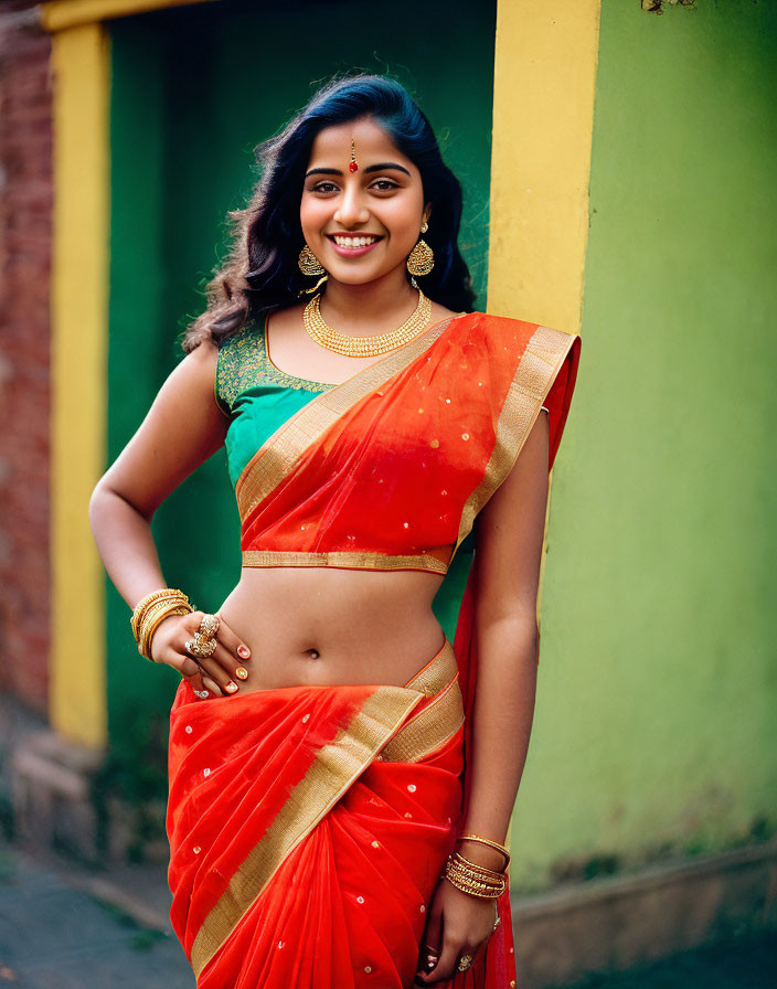 Smiling woman in red saree with gold jewelry against yellow and green wall