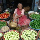 Elderly Woman in Colorful Dress Surrounded by Fresh Fruits and Vegetables at Outdoor Market