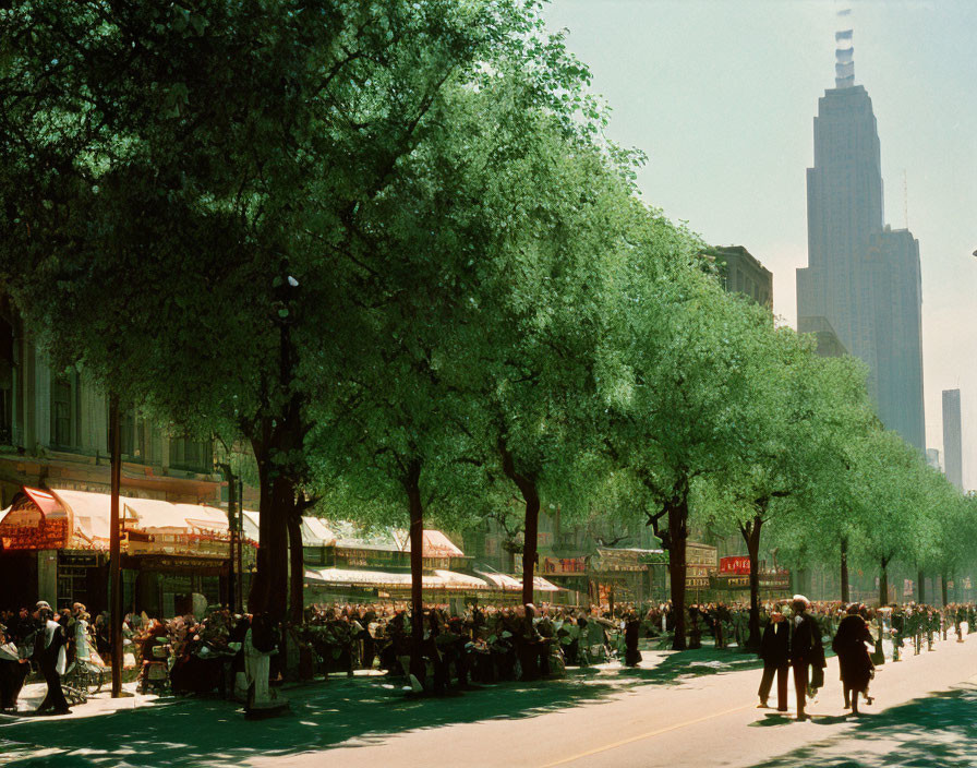 City street with trees, outdoor seating, pedestrians, and skyscraper in background