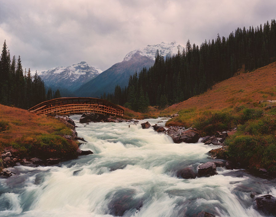 Scenic wooden bridge over rushing river with snowy mountains, pine trees, and overcast sky