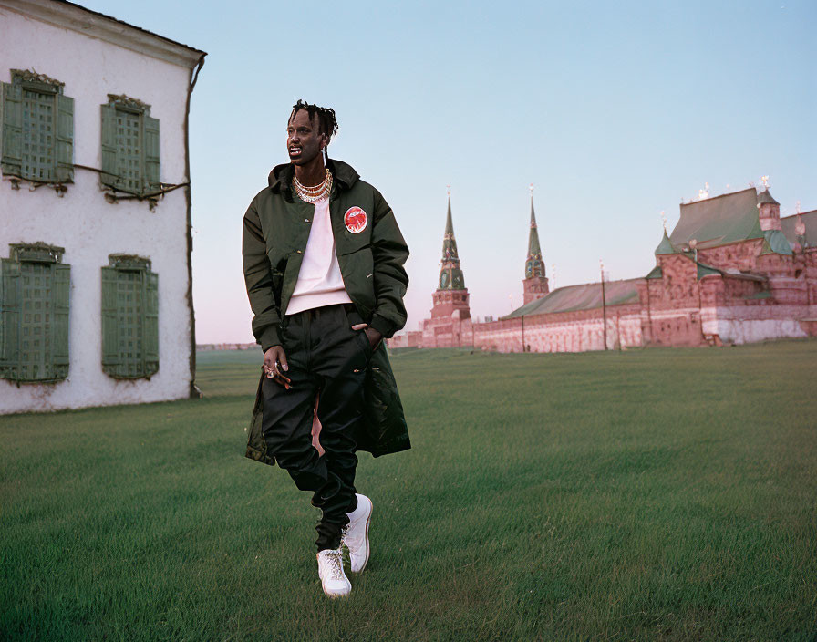 Trendy man with braided hair poses on grass near historic building at dusk