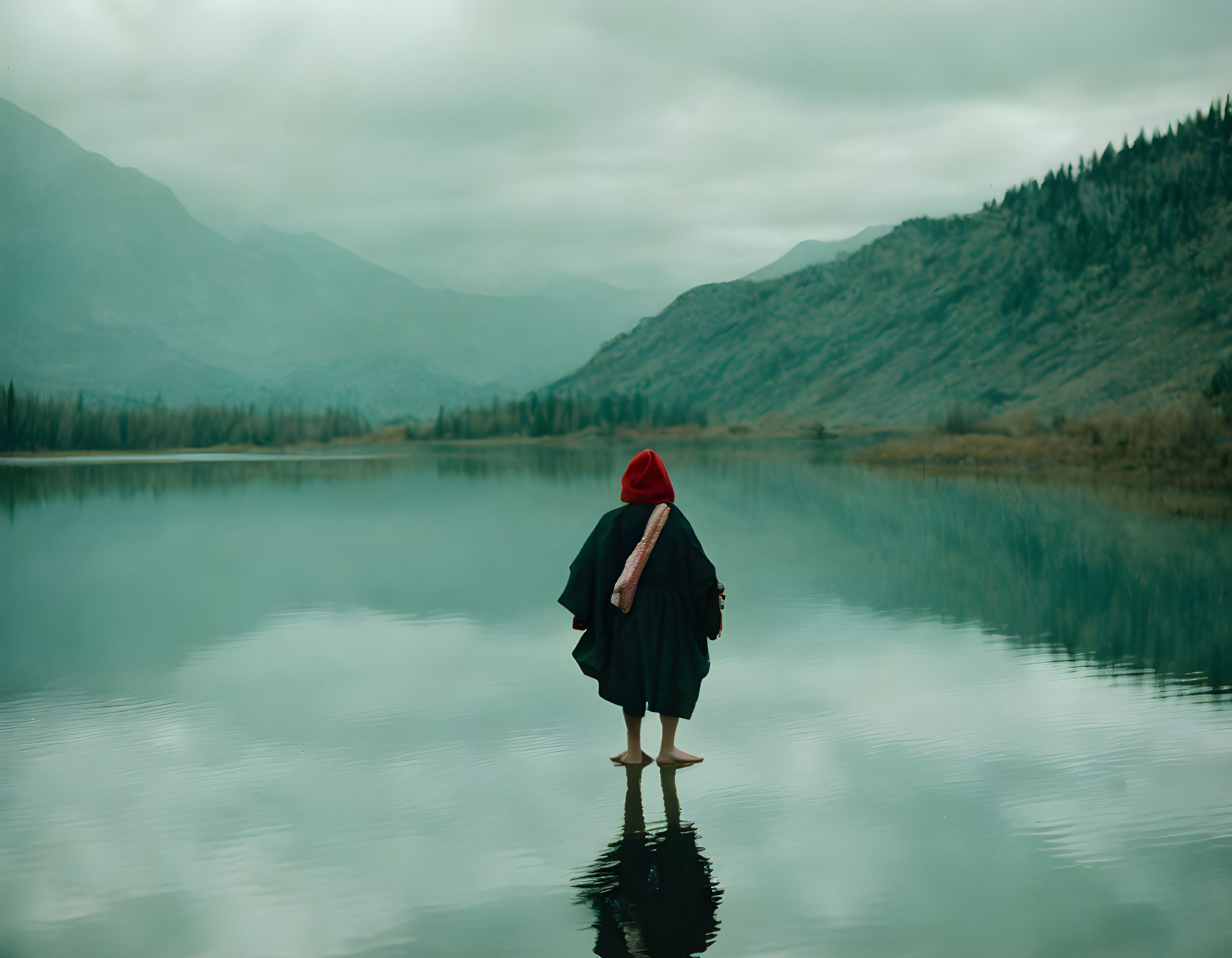 Person in Red Hat and Black Cloak Standing in Tranquil Lake with Misty Mountains