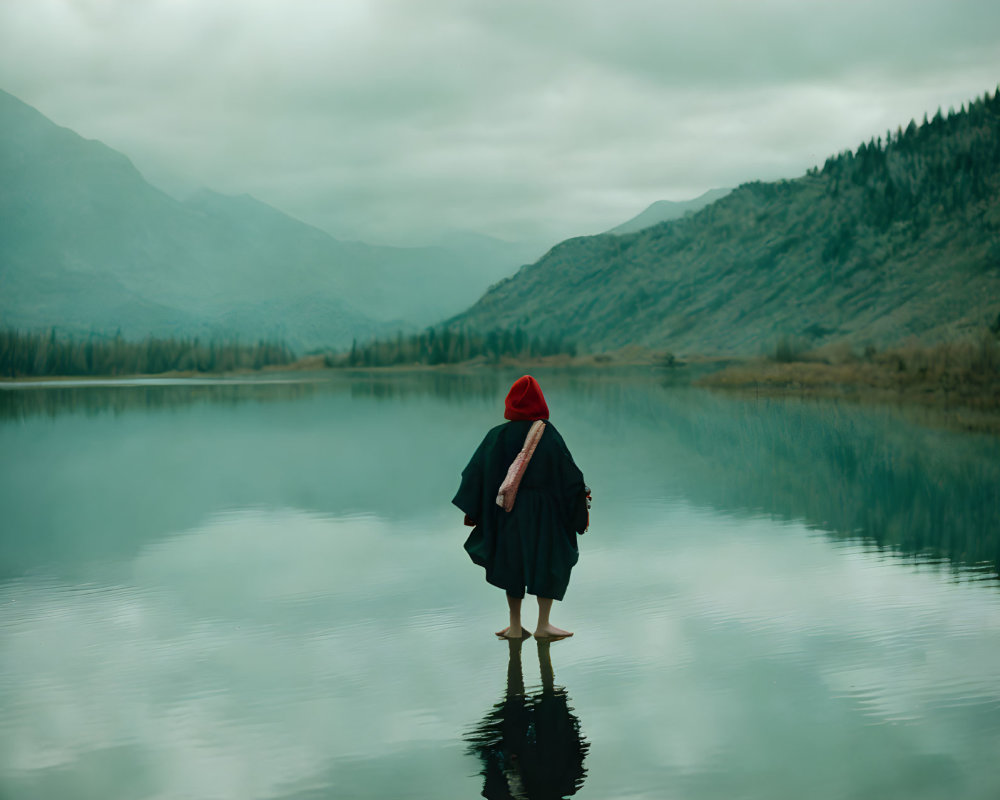 Person in Red Hat and Black Cloak Standing in Tranquil Lake with Misty Mountains