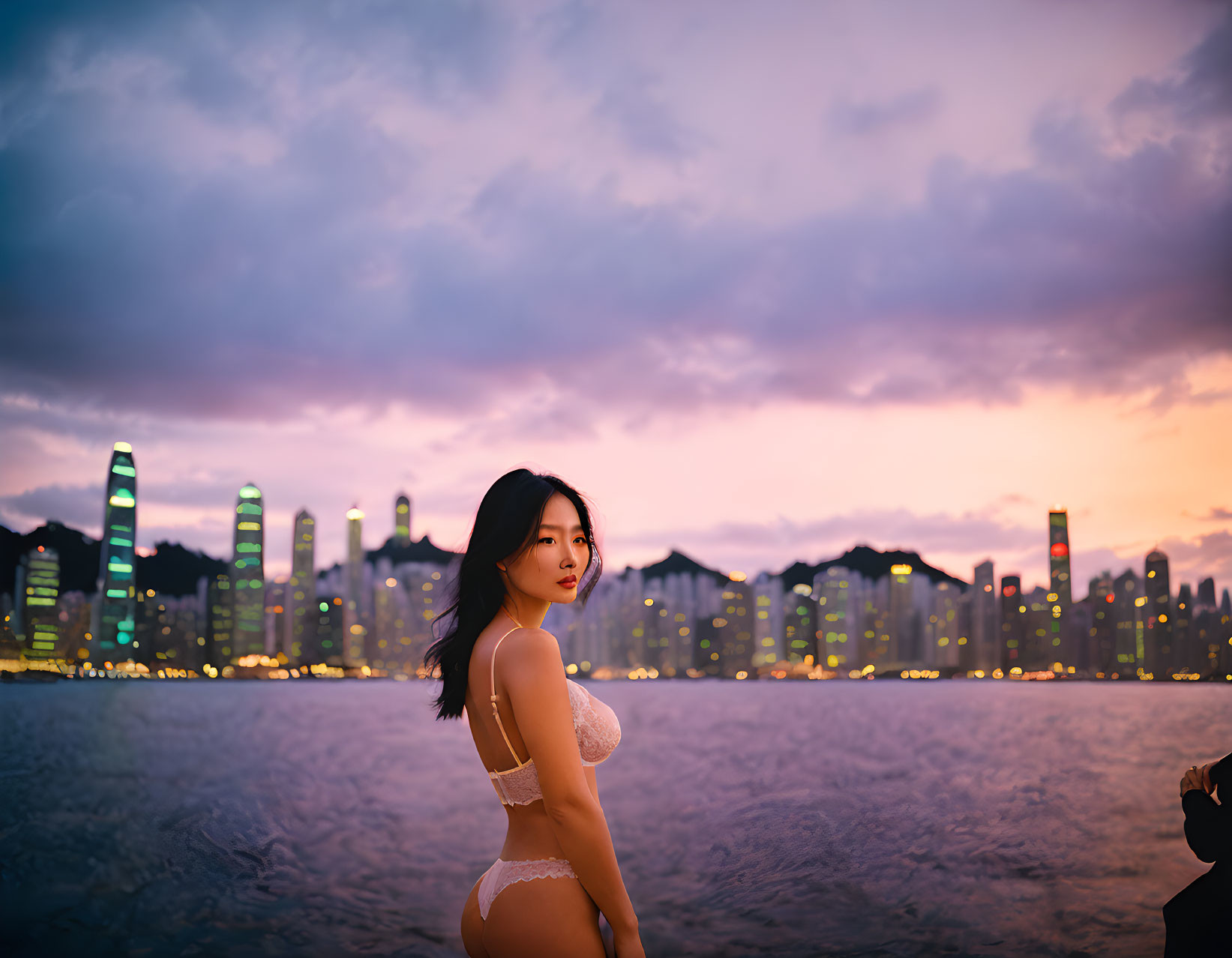 Swimwear-clad woman poses at dusk with city skyline backdrop.