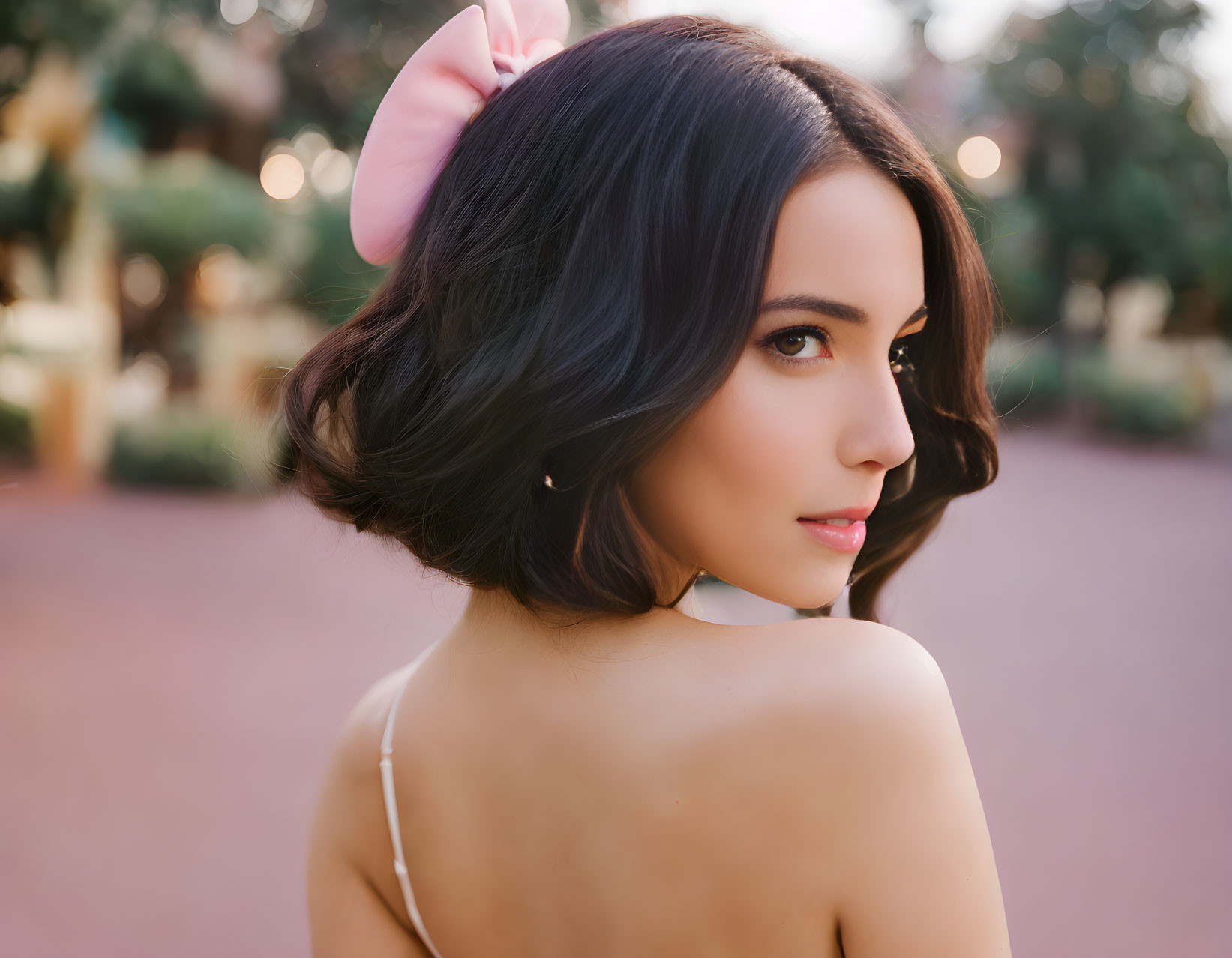 Dark-haired woman with pink hair bow and earrings in garden setting.