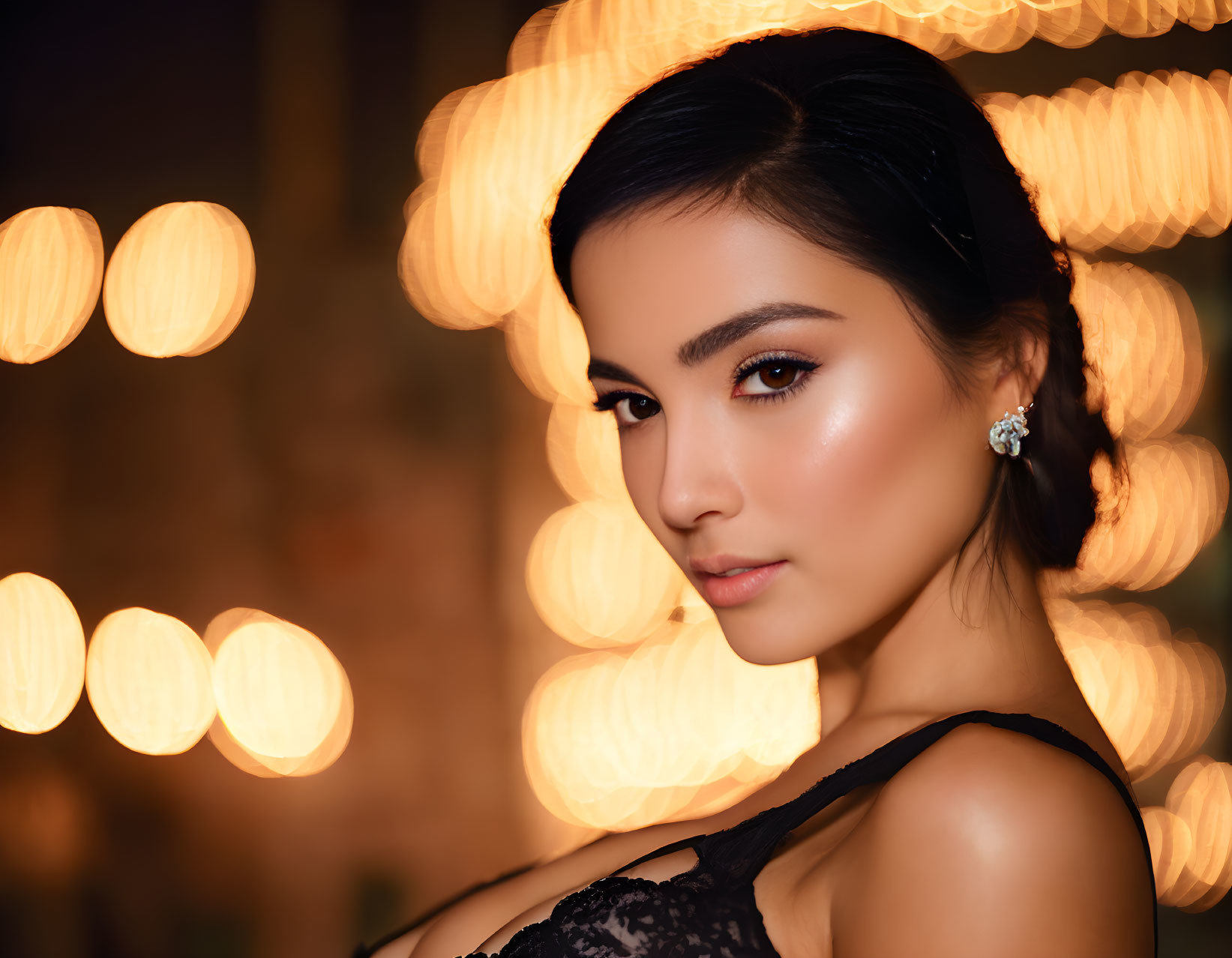 Dark-haired woman in black outfit against bokeh light background with subtle makeup and earrings.