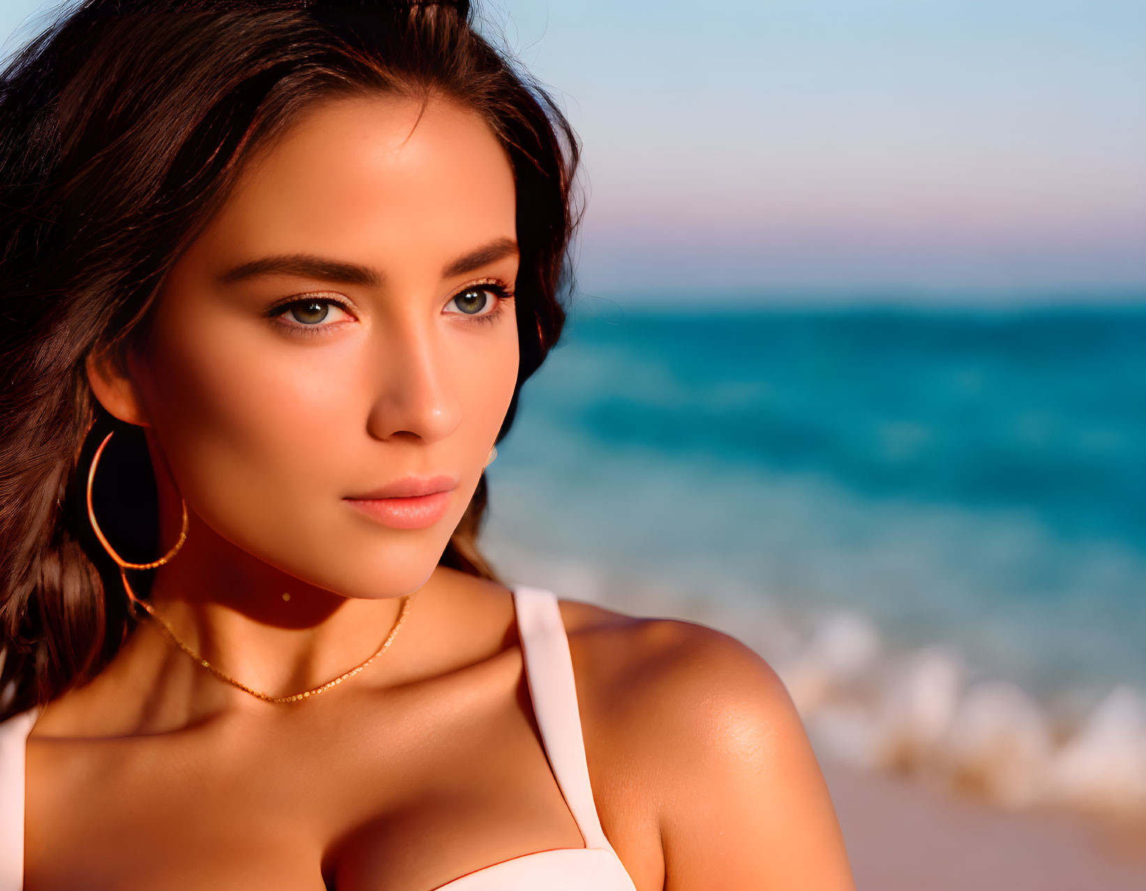 Woman with shoulder-length hair in hoop earrings and white top on beach at sunset