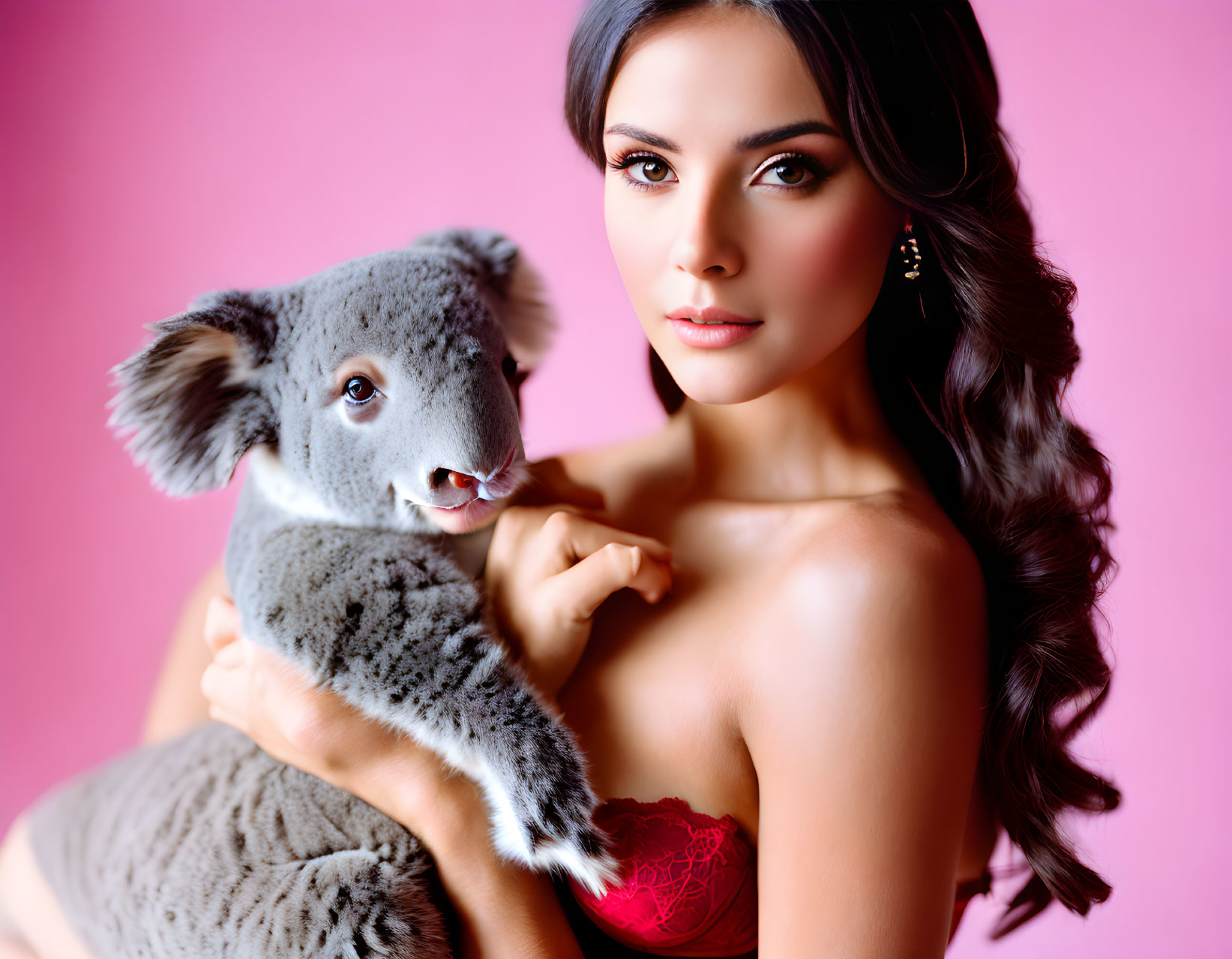 Dark-haired woman holding a gray koala on pink background with red top and earrings