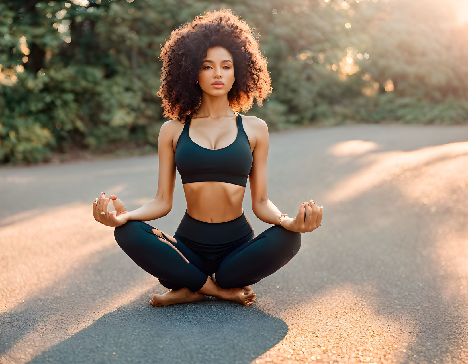 Curly-haired woman in sportswear practicing yoga in lotus position on sunlit path