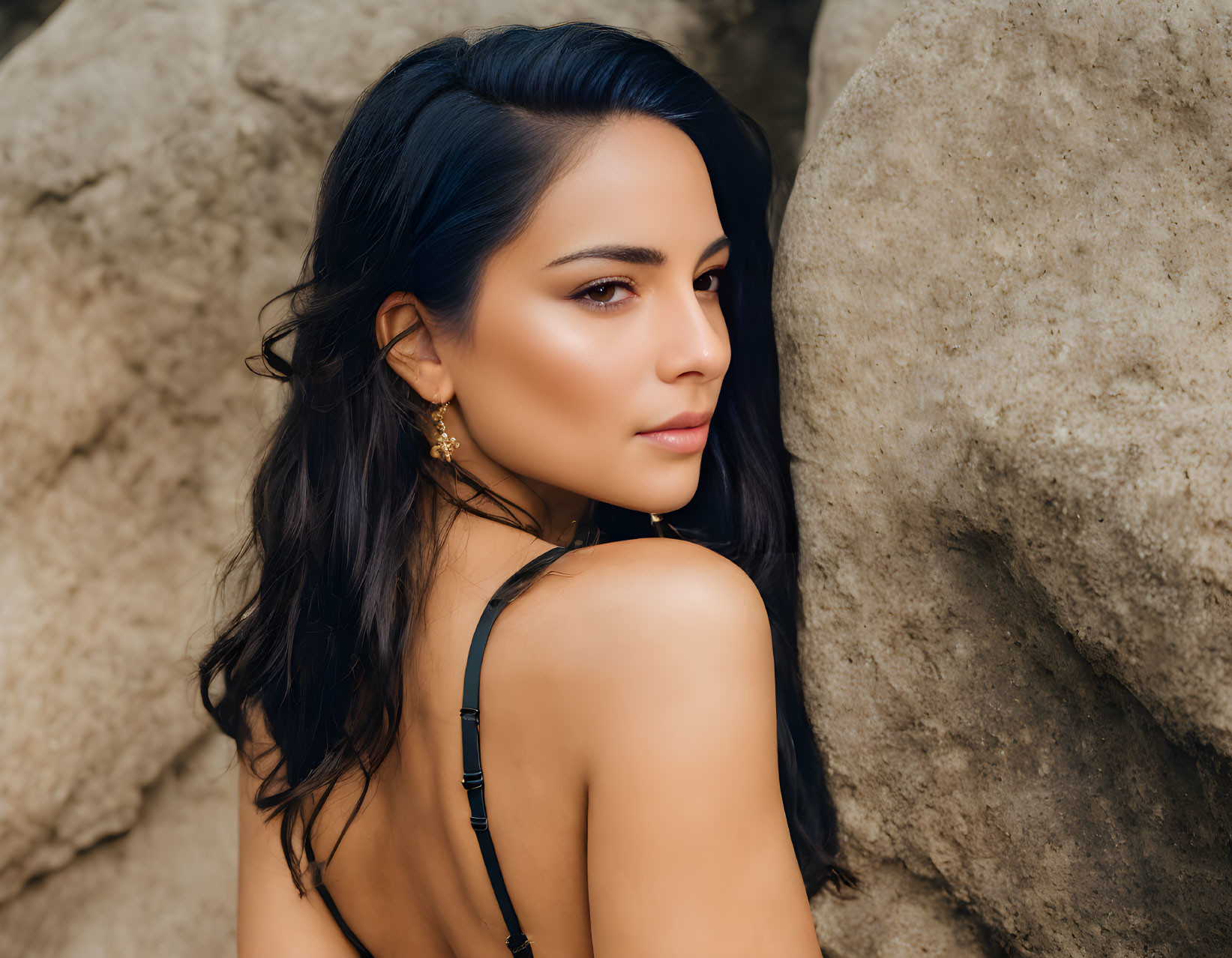 Dark-haired woman in black strappy outfit gazes over shoulder against rocky backdrop