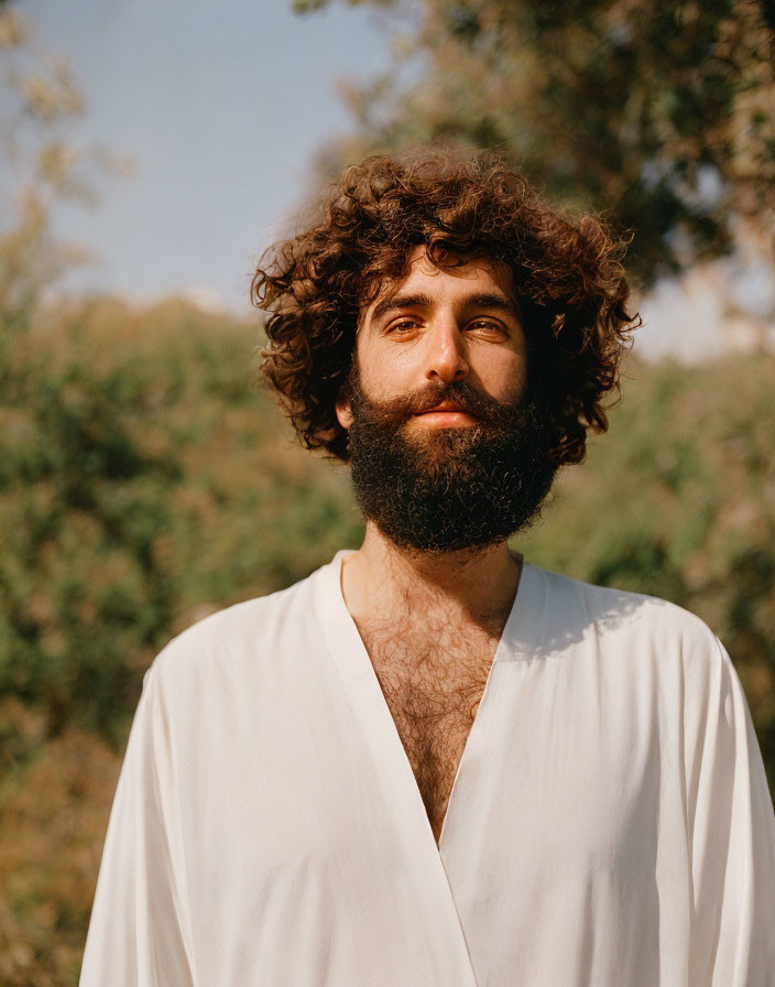 Bearded man with curly hair in white shirt smiles outdoors