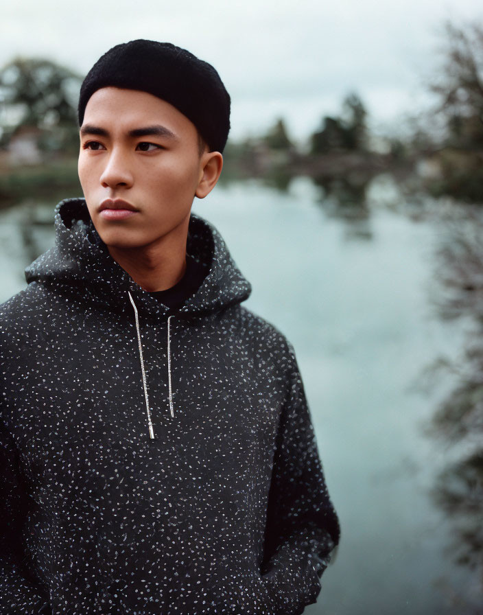 Young person in speckled hoodie and black beanie by tranquil water and trees