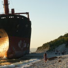 Abandoned shipwreck on rocky shore at sunset, with crashing waves and lush green hill in background