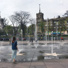 Child standing by fountain in sunny park with people and buildings