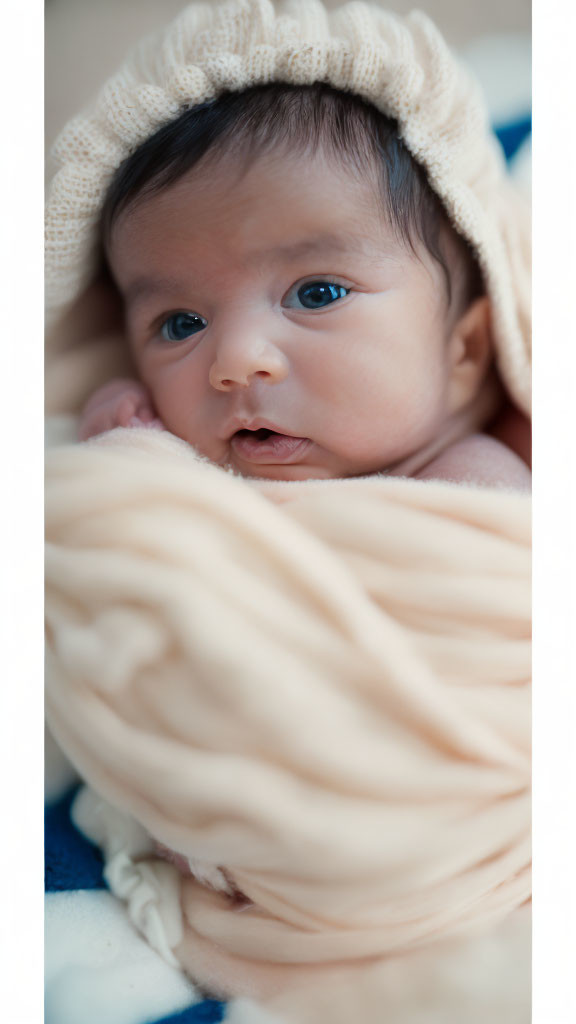 Newborn baby with dark hair in peach blanket and cream hat gazes wide-eyed