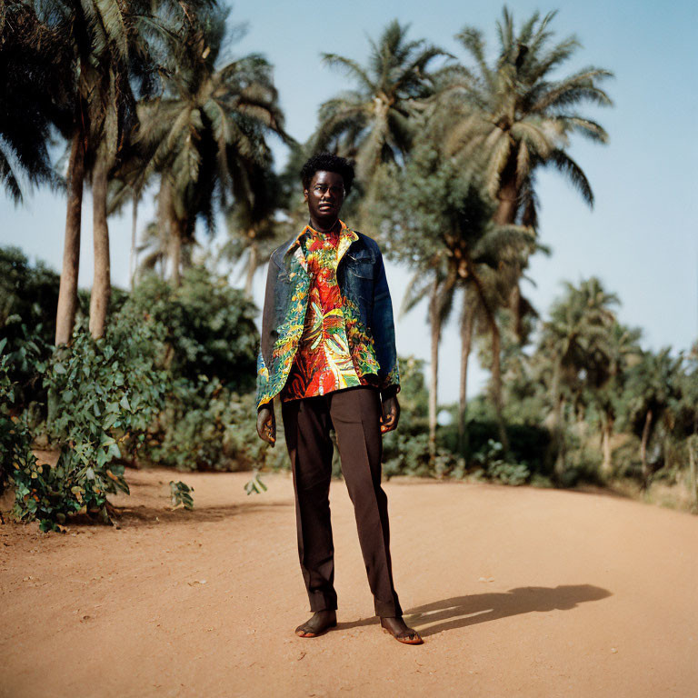 Man standing on dirt path with palm trees, wearing denim jacket and colorful shirt.