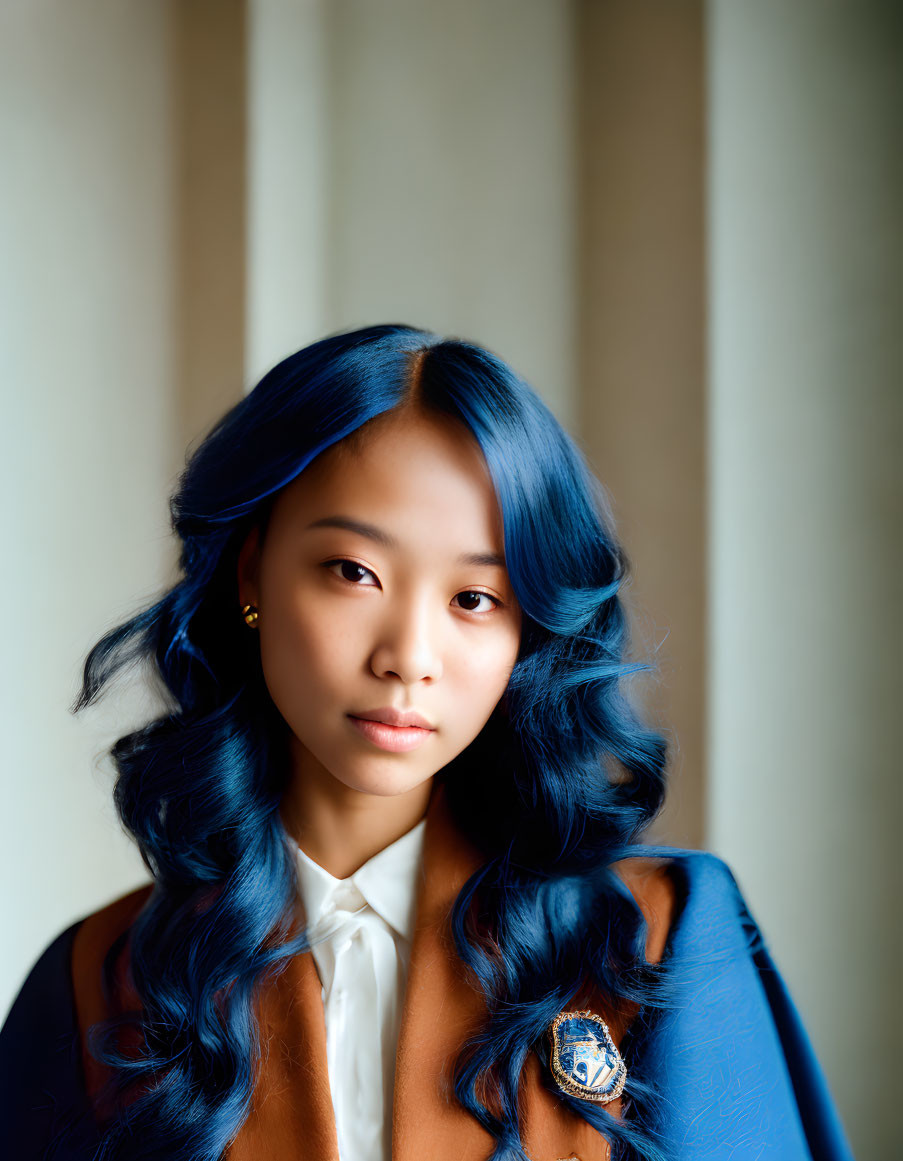 Young woman with blue hair in white blouse and brown blazer against curtain.