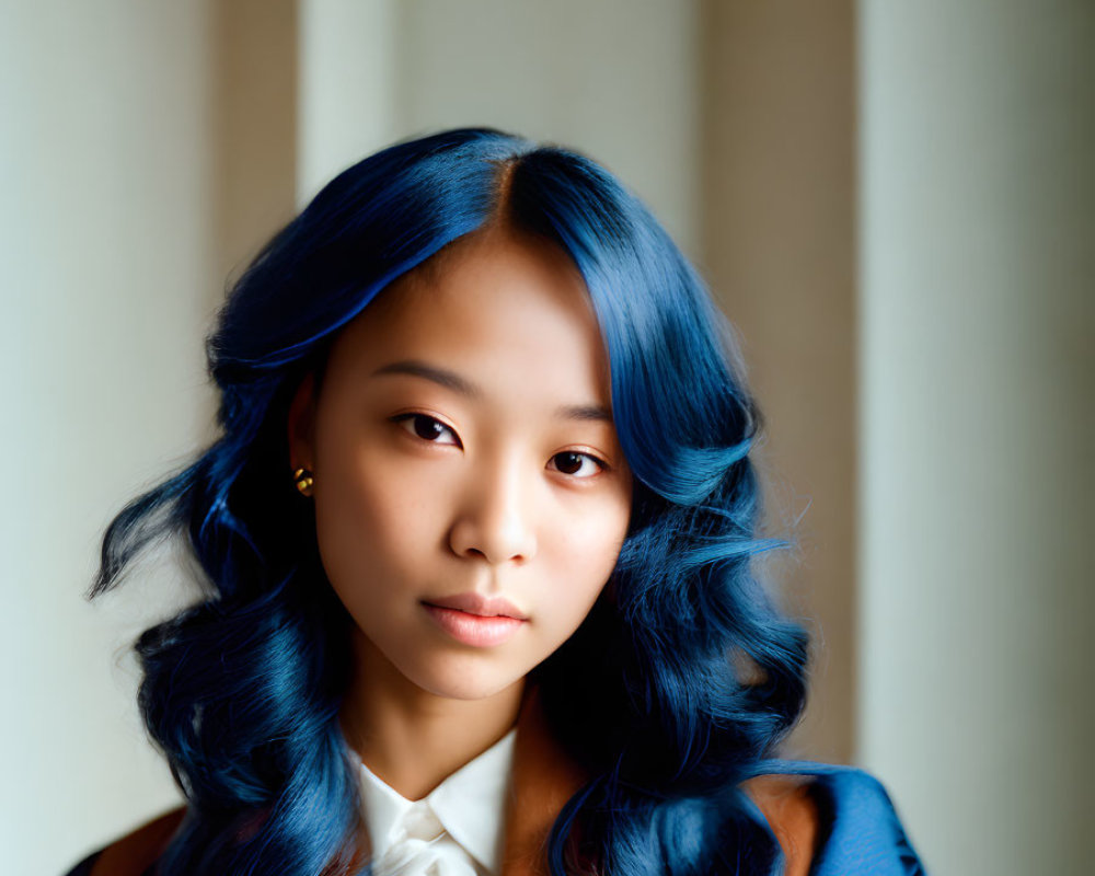 Young woman with blue hair in white blouse and brown blazer against curtain.