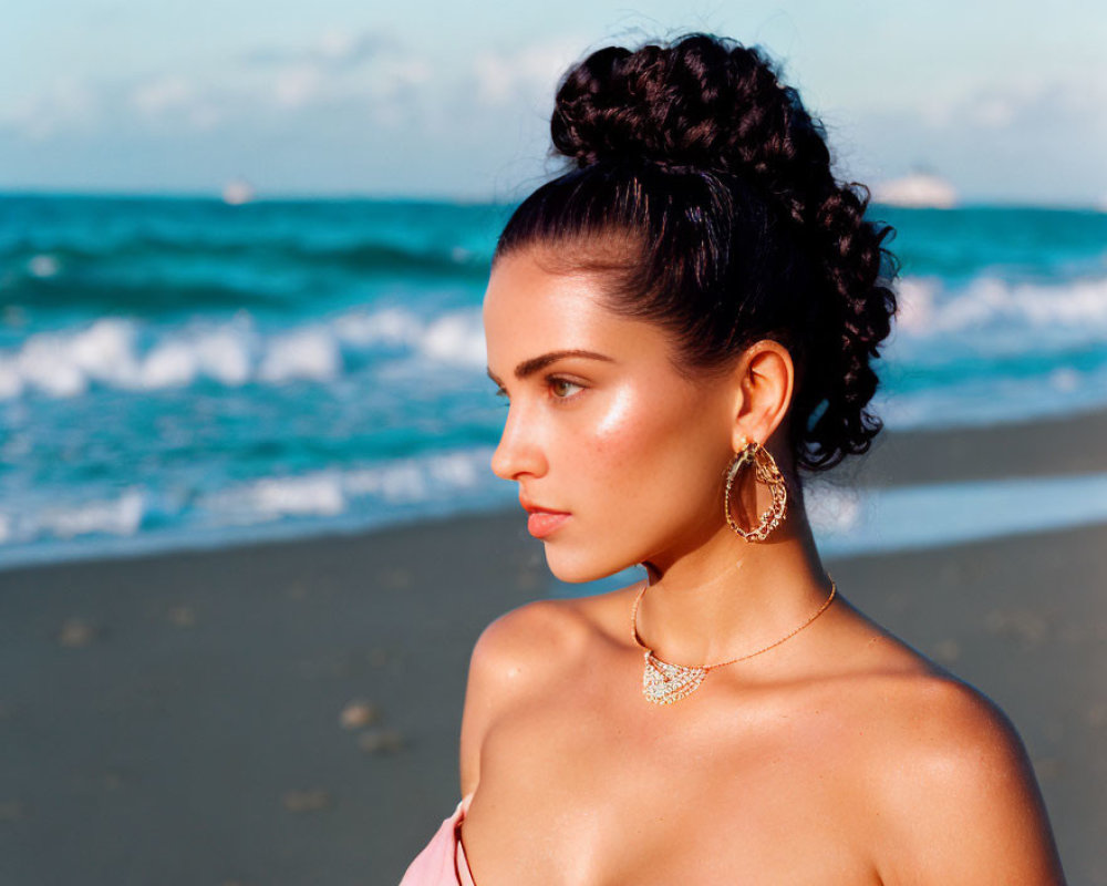 Woman with bun hairstyle at beach with earrings and necklace against ocean and blue sky.