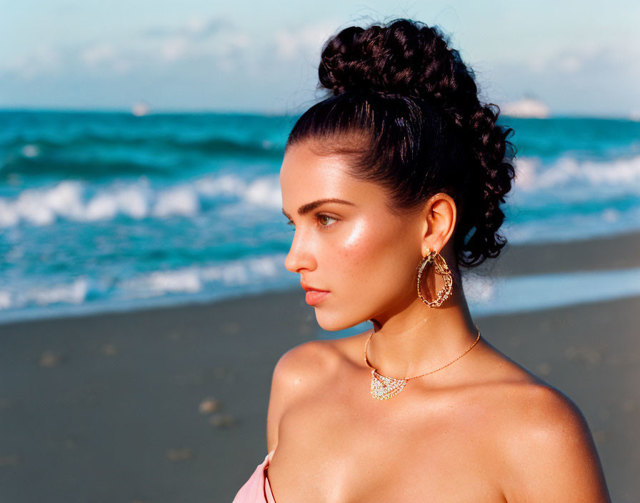 Woman with bun hairstyle at beach with earrings and necklace against ocean and blue sky.