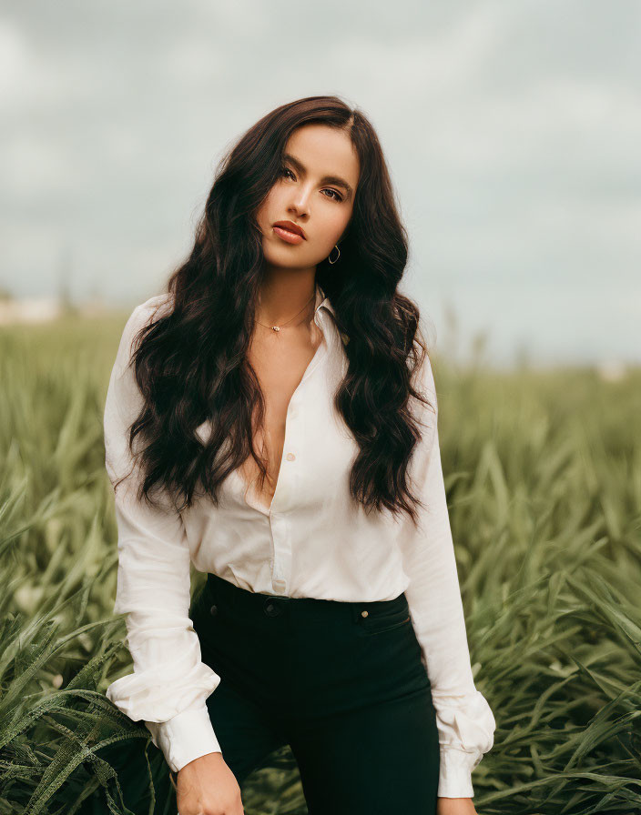 Woman with long dark hair in white blouse standing in tall grass gazes thoughtfully.