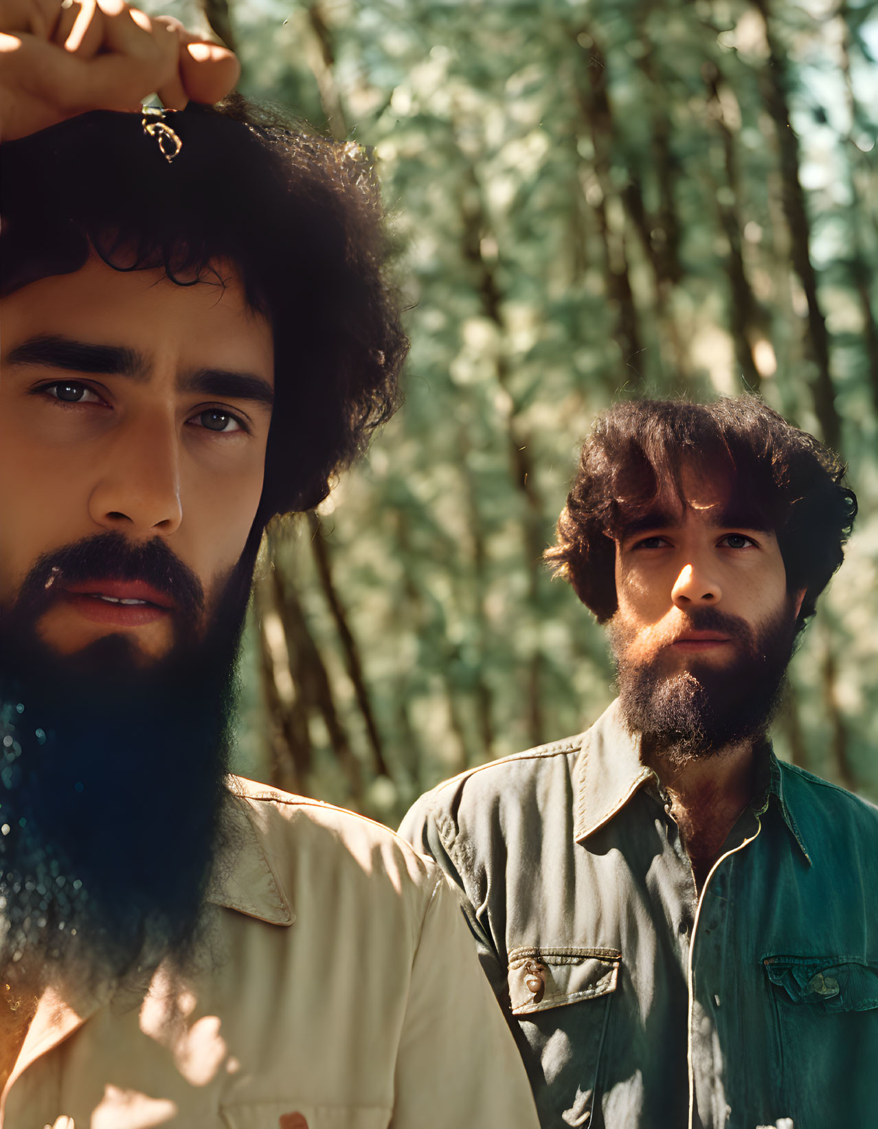 Bearded men in forest, one with contemplative expression holding small object, other blurred in background.