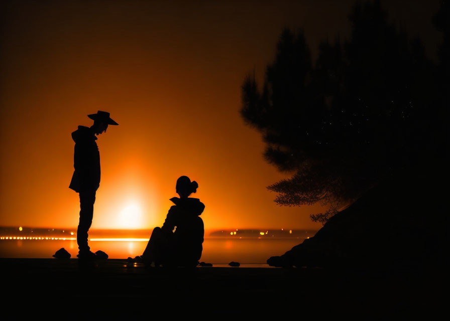 Two people silhouetted at sunset by water with tree.