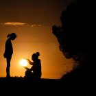 Two people silhouetted at sunset by water with tree.