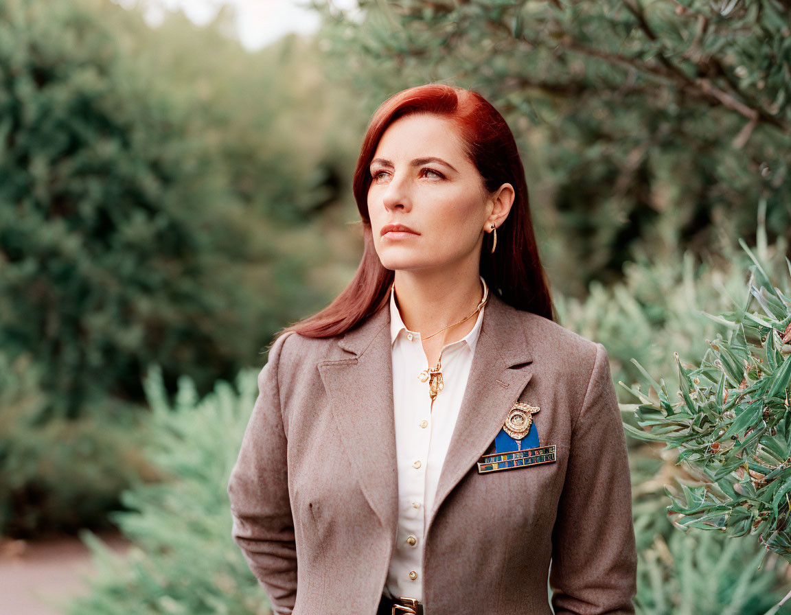 Red-Haired Woman in Blazer and White Shirt with Necklace and Badge against Greenery