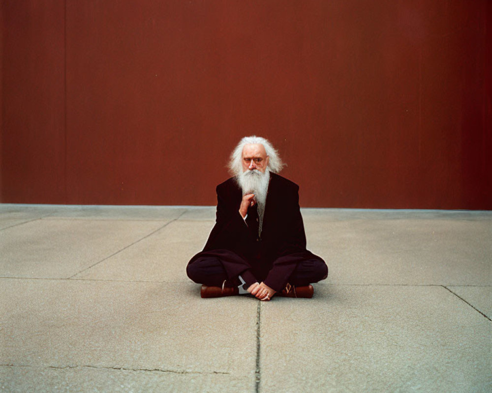 Elderly man with white hair and glasses sitting against red wall