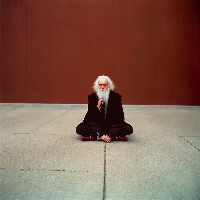 Elderly man with white hair and glasses sitting against red wall