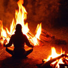 Person sitting by bright campfire at night with camping gear silhouettes