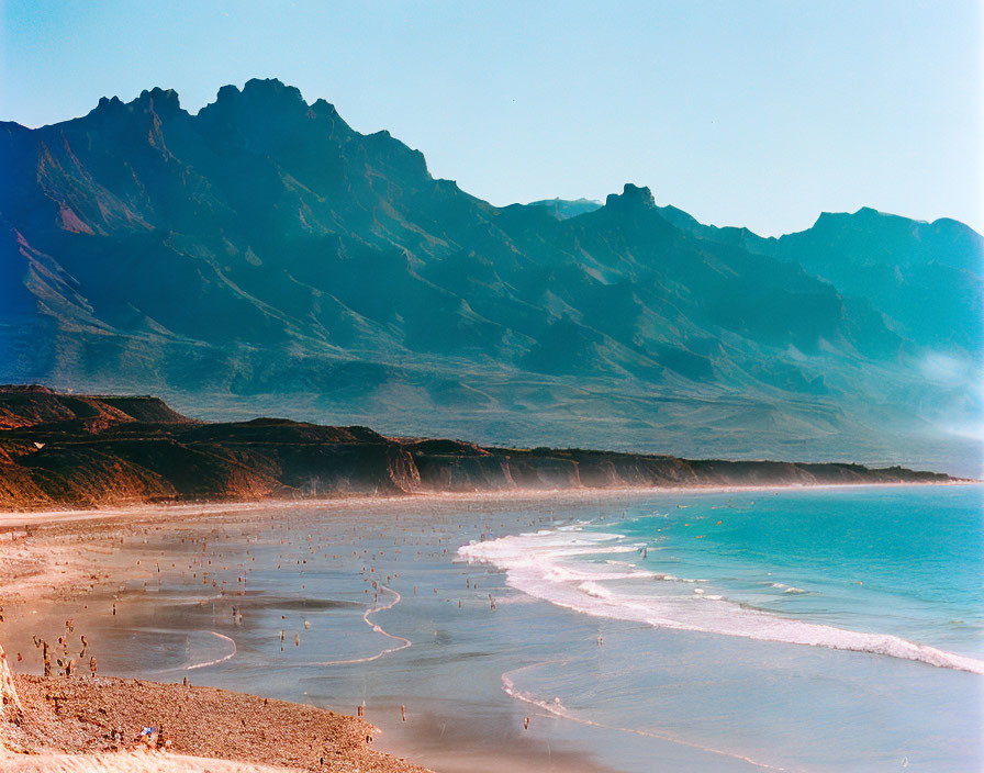 Picturesque coastline with sandy beach, gentle waves, and majestic mountain range.