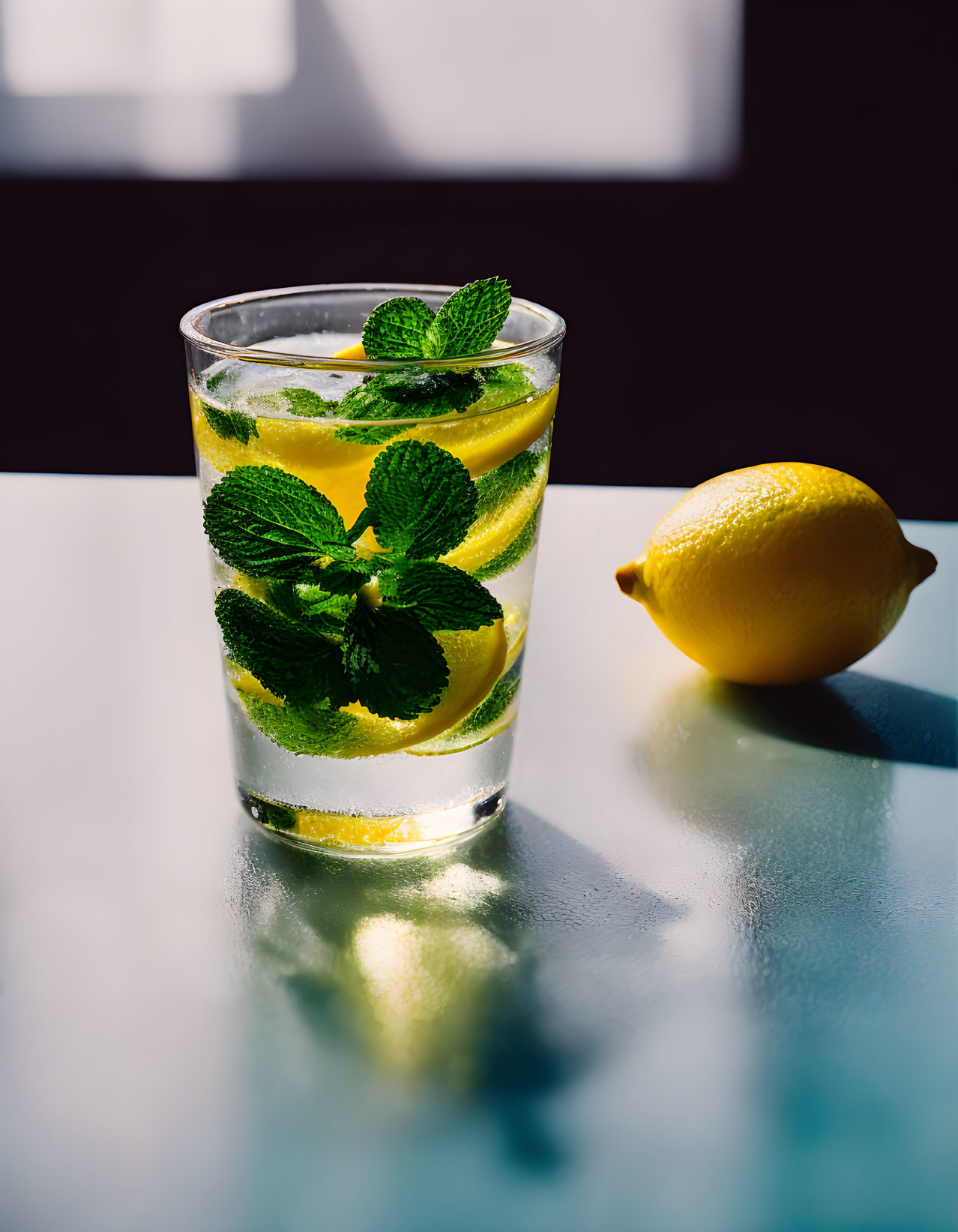 Refreshing lemon water with mint leaves and a whole lemon on reflective surface.