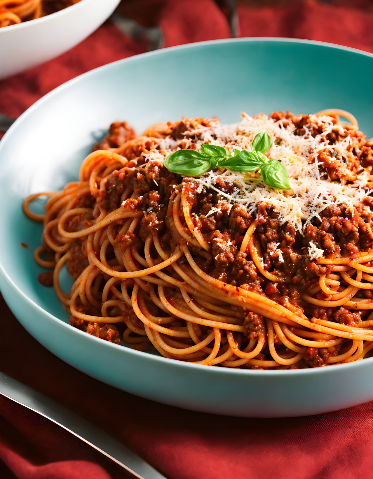 Plate of spaghetti with bolognese sauce, cheese, and basil leaf on red cloth.