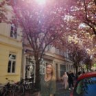 Woman walking down tree-lined path with pink blossoming trees and figure in distance