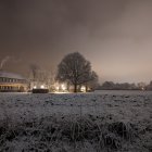 Snow-covered wintry twilight scene with bare trees and warm lights glowing from distant houses.