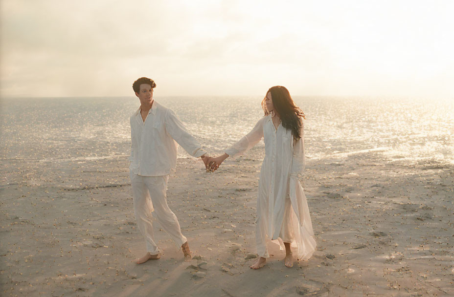 Couple holding hands on beach at sunset in white attire.