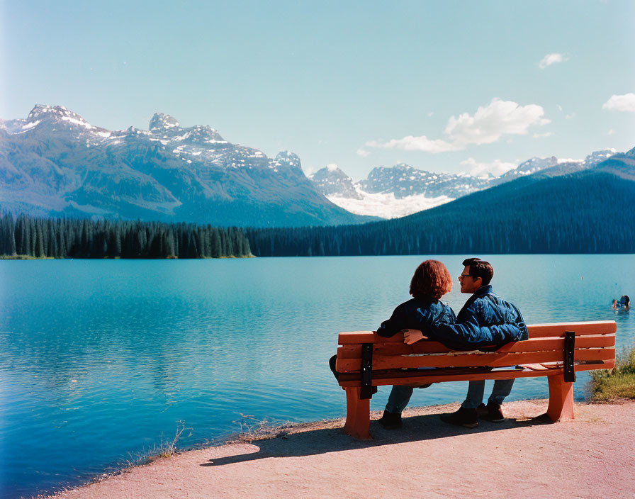 Scenic lake view with two people on bench, mountains, and forest.