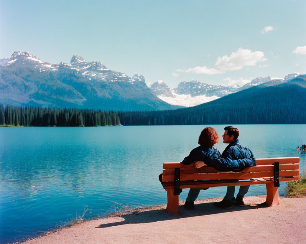 Scenic lake view with two people on bench, mountains, and forest.